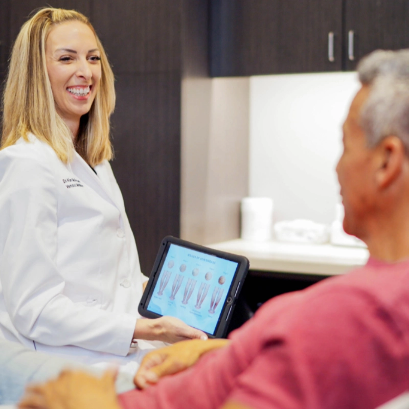 DrMcFarland_bio_page_optimized Dr. Kimberly McFarland consults with a male patient in a clinical office, using a tablet to display a diagram of the stages of vein disease. She explains the condition to the patient as part of his medical evaluation.