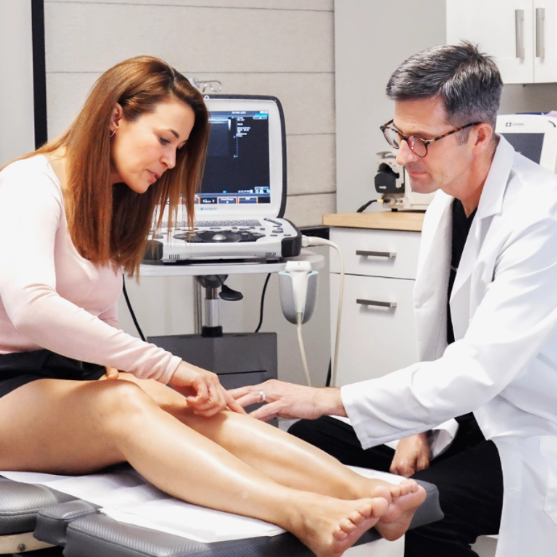 Dr. Joshua Kindelan performs a clinical consultation with a patient in an examination room. The patient points to an area on her leg while Dr. Kindelan examines it, with an ultrasound machine visible in the background.