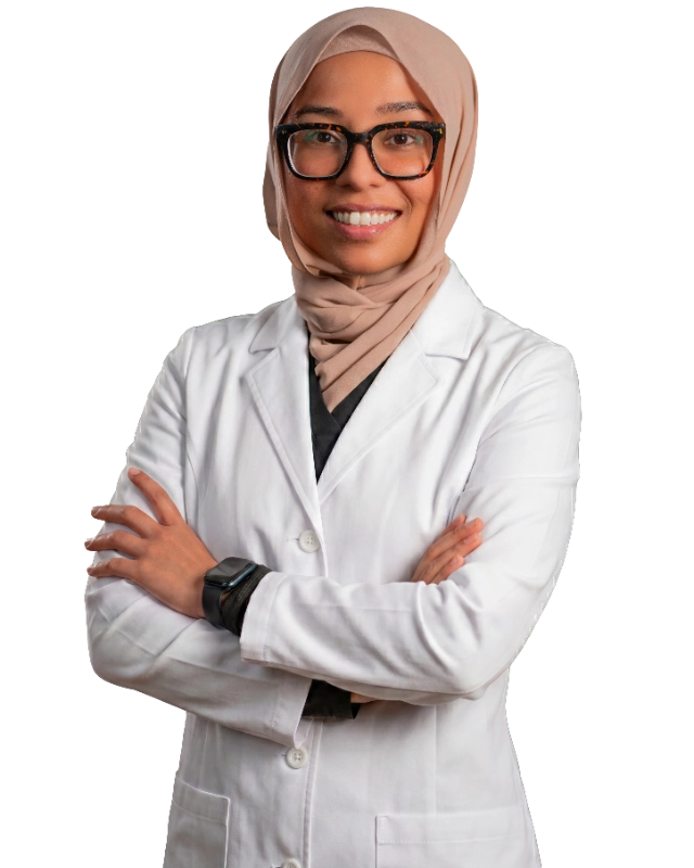 Professional headshot of Dr. Anee Khan wearing a white lab coat and black-framed glasses, smiling confidently with arms crossed. The subject is wearing a beige hijab and a smartwatch, photographed against a black background in a typical professional medical staff portrait style.