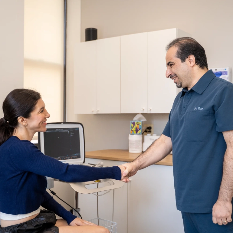 Dr. Mohsen Basiri shakes hands with a female patient in a clinical examination room, greeting or concluding a consultation. The modern medical office setting includes an Ultrasound Machine, computer workstation, and anatomical diagrams on the walls, typical of a venous or interventional pain management practice.